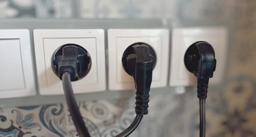 Zdjęcie przedstawia trzy czarne wtyczki podłączone do białych gniazdek elektrycznych w ścianie.
